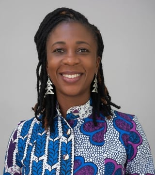 Portrait of a smiling woman with dreadlocks wearing a colorful patterned shirt and triangular earrings against a gray background